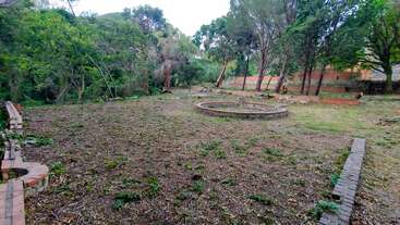 A spacious backyard with scattered grass and plants. Brick-edged garden beds and a circular raised structure are visible, surrounded by trees and bordered by a brick wall.