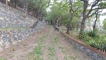 A stone pathway winds uphill between sturdy stone walls, lined with tall trees and greenery, leading through a peaceful, shaded, and natural park-like setting.