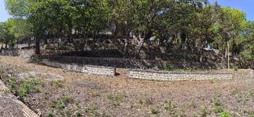 This image shows terraced stone retaining walls on a hillside with scattered trees providing shade above. The ground is dry with small patches of green vegetation.