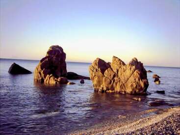 Large rocks rise from the sea near a pebbled shore under a clear sky at sunset, casting golden hues and gentle reflections on calm water.