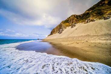 A serene beach with gentle waves touches golden sand. Steep, sandy cliffs rise dramatically to the right, under a partly cloudy sky, creating a peaceful coastal scene.