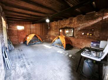 The image shows two orange tents set up inside a rustic room with brick walls, a wooden ceiling, foldable chairs, and a small cooking station with a gas stove.