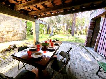 A rustic outdoor dining area with a wooden table set for six, under a wooden roof, overlooking a sunlit, green garden surrounded by trees. Inviting atmosphere.