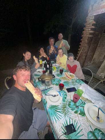 Seven people sit around an outdoor table at night, smiling and eating baguette sandwiches. The table is decorated with a tropical tablecloth and food, drinks.