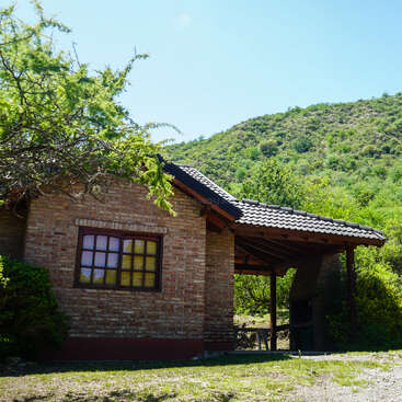 Une cabane rustique en briques, au toit de tuiles, se dresse au milieu d'une végétation luxuriante et de collines. La lumière du soleil filtre à travers les arbres, créant une atmosphère paisible et naturelle, digne d'un refuge de montagne.