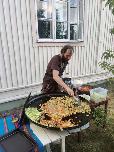 La imagen muestra a un hombre cocinando al aire libre en una gran sartén, de pie frente a una casa blanca con una ventana, rodeado de diversos ingredientes y utensilios de cocina.
