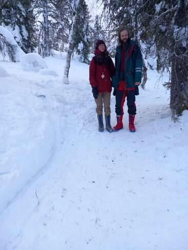 La imagen representa una serena escena invernal en la que aparecen un hombre y una mujer juntos en un bosque nevado, rodeados de árboles y terreno nevado.