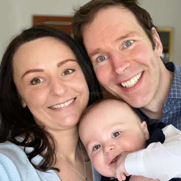 The image depicts a family of three, with a woman, a man, and an infant, posing for a selfie, showcasing their affectionate bond.