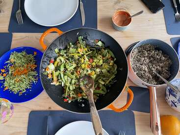 The image depicts a table with a variety of dishes, including a large wok filled with vegetables, a bowl of rice, and a plate of salad, set for a meal.