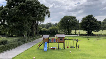 The image depicts a wooden play structure with a slide and climbing bars, surrounded by a grassy area, featuring a wooden fence and a tree in the background. The structure is situated in a fenced yard.