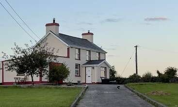 The image depicts a large house with a gable roof, featuring a prominent front door and a row of windows, set against a backdrop of a well-manicured lawn and a clear sky.