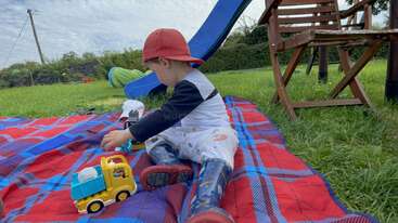 A young boy is playing outside with a toy truck, a ball, and a frisbee. He is wearing a t-shirt and shorts and is standing on a grassy area. The background is a park or a field.