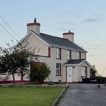 The image depicts a large house with a gable roof, featuring a prominent front door and a row of windows, set against a backdrop of a well-manicured lawn and a clear sky.