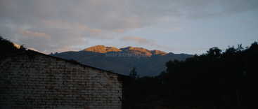Uma casa rústica de tijolos ergue-se em primeiro plano, enquanto os picos das montanhas, banhados pela luz do sol, brilham sob um céu nublado, rodeados por árvores sombrias à medida que o anoitecer se instala suavemente.