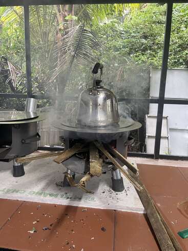 Une installation traditionnelle de cuisine en plein air avec une bouilloire en métal chauffée sur un poêle à bois. La fumée s'élève, entourée d'une végétation luxuriante et de carreaux rustiques, créant une atmosphère chaleureuse.