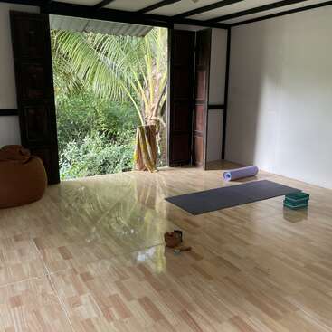 Un espace de yoga paisible avec de grandes portes ouvertes, un tapis de yoga, des blocs, un bolster et un pouf, avec vue sur une végétation luxuriante et un palmier à l'extérieur.