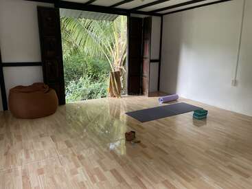 A peaceful yoga space with large open doors, a yoga mat, blocks, bolster, and bean bag, overlooking lush greenery and a palm tree outside.