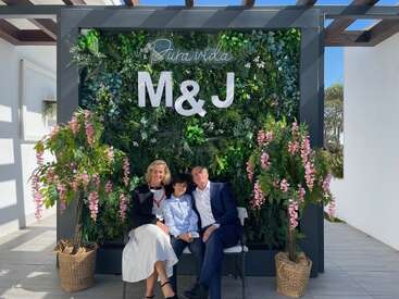 Três pessoas sentadas juntas, sorrindo em frente a uma parede verde decorada com flores, letras grandes “M & J” e a frase “Pura vida” no topo.