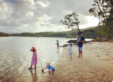 Uma família se diverte pescando na margem arenosa de um rio, sob um céu nublado, cercada por árvores. Crianças vestidas com roupas coloridas brincam e exploram perto da beira da água.