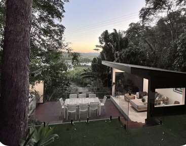 Esta imagem mostra um pátio moderno ao ar livre com uma longa mesa de jantar, cercado por árvores frondosas, ao lado de uma elegante sala de estar aberta ao pôr do sol.