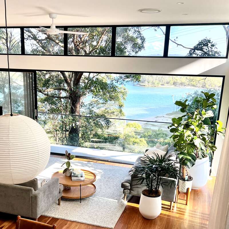 A bright modern living room with large windows overlooking a serene lake, lush greenery, comfortable furniture, stylish decor, and vibrant indoor plants create a tranquil atmosphere.