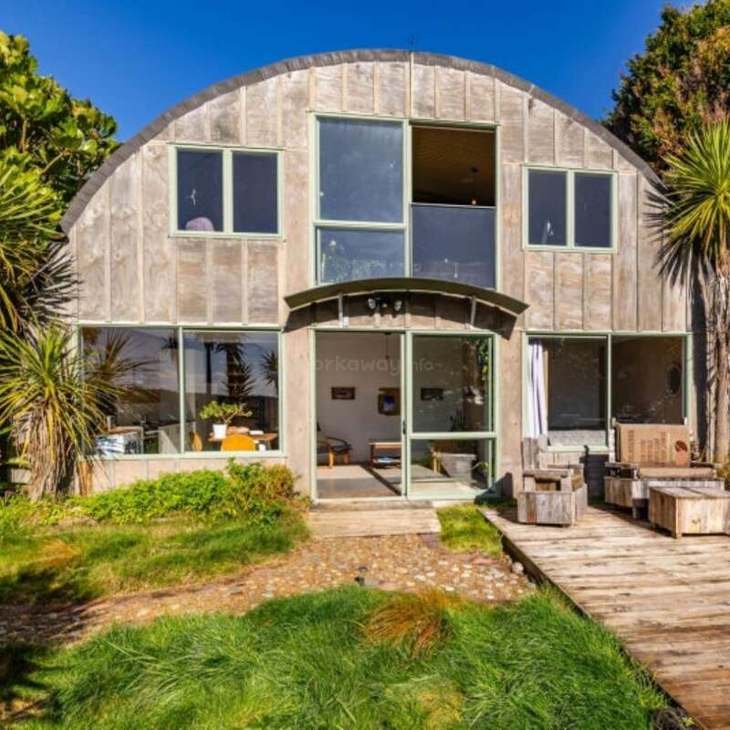 A imagem mostra uma casa moderna com um design exclusivo, com um telhado plano e uma grande janela, tendo como pano de fundo um espaço externo sereno. A casa é cercada por um gramado bem cuidado.