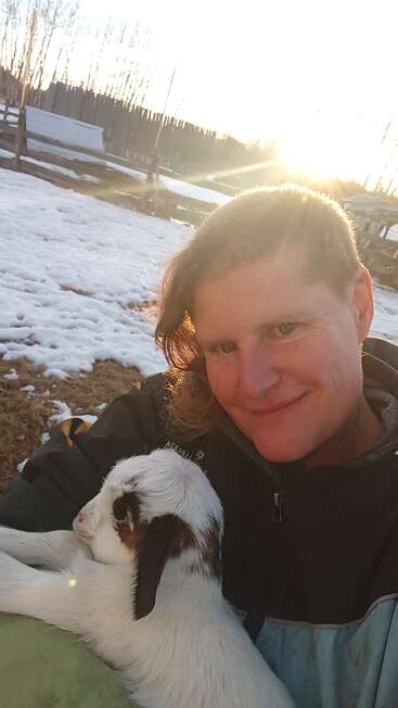 A smiling person holds a small white goat with brown spots. Snow and patches of grass cover the ground, while sunlight streams in the background. Peaceful moment.