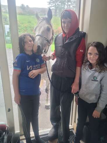 Three children stand inside by a door, accompanied by a horse poking its head in. They all appear calm, with a green landscape visible outside.