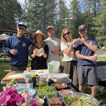 Five friends pose outdoors at a picnic table covered in colorful food and salad bowls. Tall trees surround them, creating a cheerful, sunny, forest gathering.