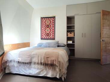 Um quarto aconchegante com a cama bem arrumada, tons neutros, manta de tecido, almofadas, tapeçaria étnica na parede, guarda-roupa embutido, prateleiras, cesta, porta aberta e luz natural.