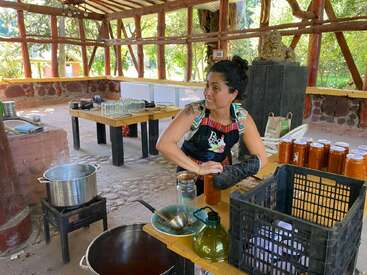 Uma mulher está engarrafando conservas caseiras numa cozinha rústica ao ar livre. Ela está usando um avental e uma luva de forno, cercada por potes, panelas e uma paisagem natural.