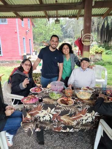 Cinco pessoas estão reunidas ao ar livre em torno de uma mesa cheia de comida, sorrindo e curtindo uma refeição juntas. O ambiente é descontraído, sob um telhado, perto de uma casa vermelha.