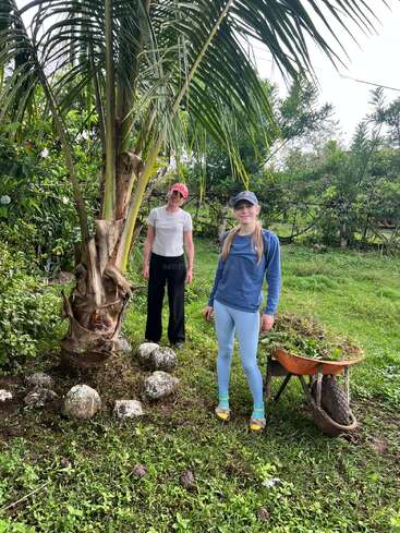 Dos personas se encuentran junto a una palmera en un frondoso jardín. Una de ellas sostiene una carretilla llena de plantas. Ambos llevan sombreros y ropa informal de exterior, y sonríen.