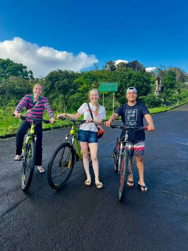 Tres amigos posan con sus bicicletas en una carretera asfaltada, rodeados de exuberante vegetación y un cielo azul brillante. Parecen felices y listos para la aventura.
