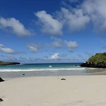 A beautiful sandy beach with clear turquoise water, surrounded by green rocky cliffs under a bright blue sky scattered with fluffy white clouds on a sunny day.