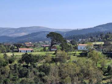 Esta cena rural serena mostra colinas onduladas, casas espalhadas, árvores verdejantes e campos sob um céu azul claro, com montanhas ao fundo. A beleza tranquila da paisagem.