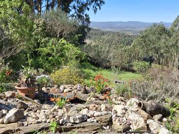 Um jardim rochoso com flores coloridas, vasos de plantas e cerâmicas quebradas fica em primeiro plano. A vegetação exuberante e as colinas arborizadas ao longe completam a paisagem.