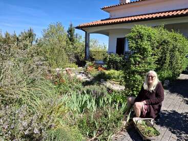 Uma mulher de cabelos loiros compridos está sentada ao lado de um jardim verdejante, segurando uma cesta de ervas. Atrás dela, há uma casa com telhado vermelho.