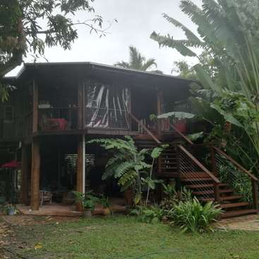 The image depicts a two-story wooden house with a balcony and stairs, surrounded by lush tropical plants and trees, set against an overcast sky.