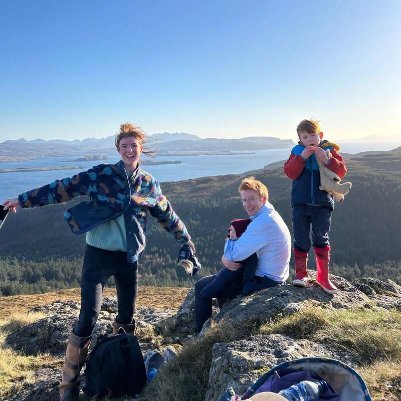 Una familia feliz disfruta del aire libre en lo alto de una pintoresca colina. Con vistas al agua y a las montañas lejanas, sonríen y juegan juntos con el tiempo fresco y soleado.