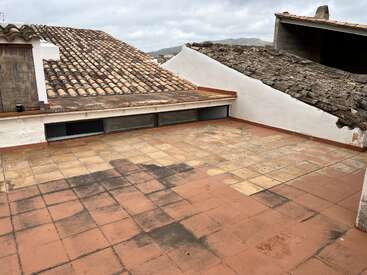 Cette image montre une terrasse sur le toit aux tuiles patinées et tachées, entourée de toits traditionnels en tuiles d'argile et de murs blancs, sous un ciel nuageux avec des collines au loin.