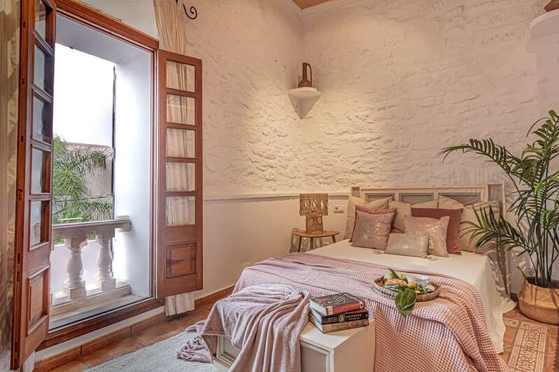 Cette chambre chaleureuse se distingue par un mur en pierre blanche au charme rustique, une porte en bois ouverte donnant sur un balcon, un lit confortable agrémenté de coussins, une plante et des livres posés sur un coffre.
