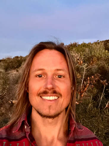 A smiling person with long hair and a mustache takes a selfie outdoors. They wear a red patterned shirt. The background includes greenery and a blue sky.