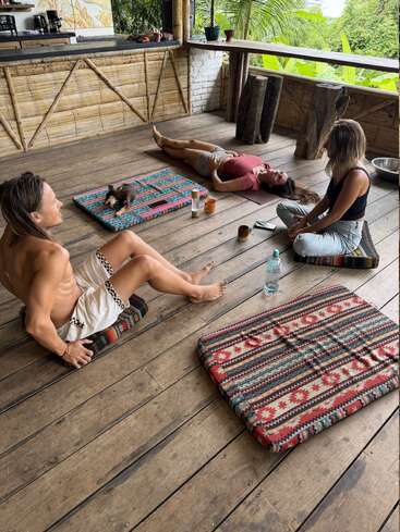 Three people relax on patterned cushions in a rustic, open-air kitchen. One lies down, a small dog rests nearby. Surrounded by greenery, mugs, and a water bottle.