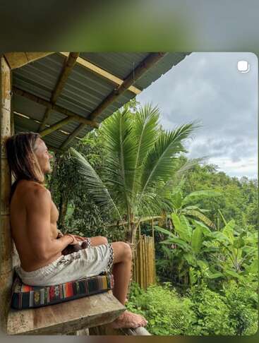A shirtless man sits peacefully on a colorful cushion on a veranda, gazing at lush tropical greenery, surrounded by palm trees and dense vibrant foliage.