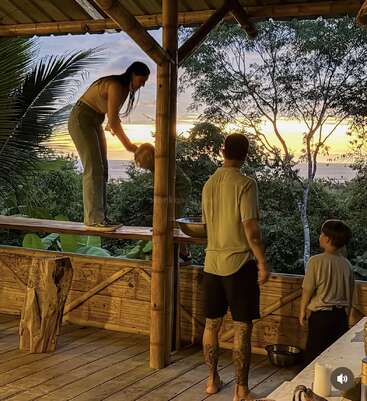 A woman stands on a wooden ledge, preparing a coconut, while a man and child watch. Surrounded by lush greenery, the sun sets beautifully in the background.