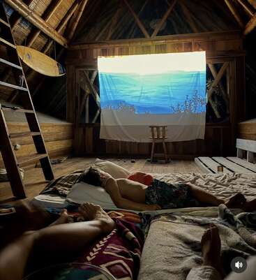 In a cozy wooden attic, two people relax on blankets, watching a projected ocean scene on a hanging sheet. A surfboard and ladder add rustic charm.