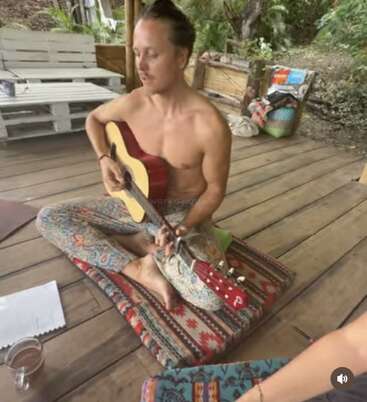 A shirtless man sits cross-legged on a patterned cushion, playing guitar on a wooden deck surrounded by nature. Papers and a drink sit nearby.