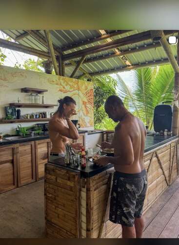 Two shirtless men are preparing food together in an open-air bamboo kitchen, laughing and enjoying the tropical surroundings with palm trees and nature visible outside.