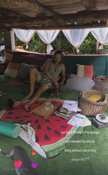 A man relaxes in a cozy jungle hut, surrounded by pillows, books, and nature. Sunlight filters through curtains; a cat and simple decor complete the tranquil scene.